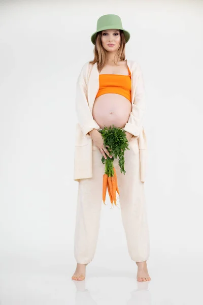 Молодая красивая беременная женщина с длинными темными волосами в зеленой шляпе ведро стоя, держа свежую кучу моркови. — стоковое фото