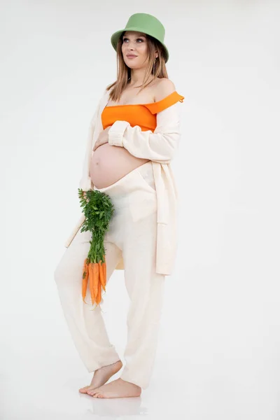 年轻美丽的孕妇，一头乌黑的长发，戴着绿色水桶帽，站着，抱着肚子和一堆胡萝卜. — 图库照片