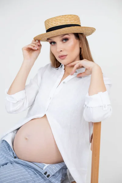 길고 검은 머리에 화장을 한 임신 한 젊은 여성의 모습은 흰 셔츠, 지퍼를 벗기지 않은 청바지와 지푸라기 모자를 착용하고 있다. — 스톡 사진