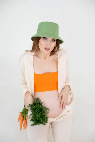年轻美丽的孕妇，一头乌黑的长发，戴着绿色水桶帽，坐着，抱着肚子和一堆胡萝卜. — 图库照片
