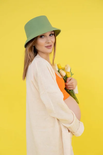 Портрет молодой милой босиком беременной женщины в зеленой шляпе ведро, оранжевый топ, стоя, держа кучу тюльпанов. — стоковое фото