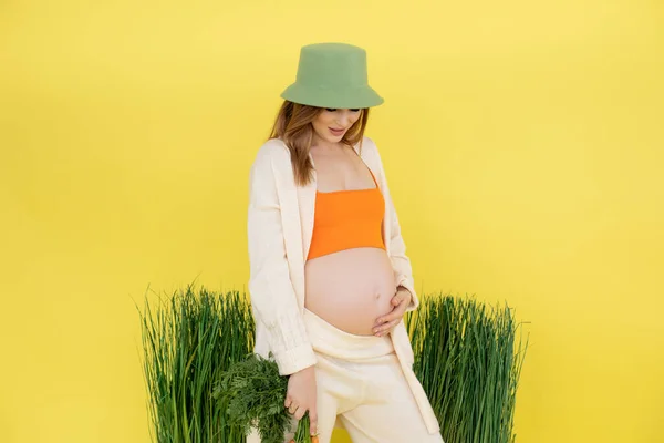 Молодая улыбающаяся красивая беременная женщина носит зеленую шляпу ведро, оранжевый топ, бежевый кардиган с кучей свежей моркови. — стоковое фото