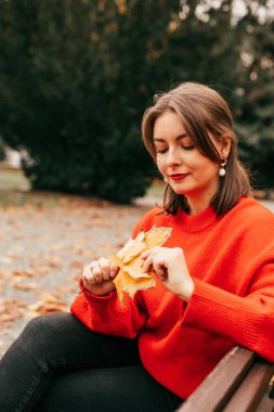 Çok düşünceli bir bayan öğrencinin dikey portresi, bankta oturuyor, akçaağaç yaprakları tutuyor. Sonbahar ağacı görünümünü park et