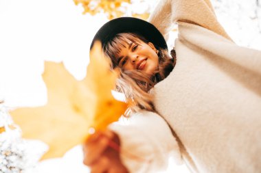 Fotoğrafta gülümseyen çekici kadın kameraya bakıyor, elinde altın akçaağaç yaprağı var. Boş zaman aktivitesi. Düşük açı