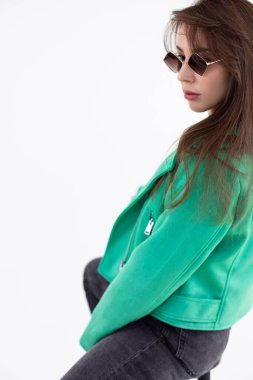 Sandalyede oturan siyah kot ve güneş gözlüklü yeşil ceketli bir kadın portresi. Moda aksesuarlı ve şık giyinen çekici bir kadın. Moda fotoğrafı, stüdyoda beyaz arka planda poz.