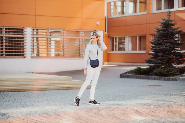 Блондинка довольно стильная женщина в солнечных очках, плечевая сумка случайная белая одежда ходьба по улице возле здания ели. — стоковое фото