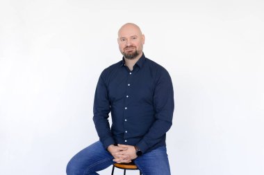 Memnun orta yaşlı bir adamın portresi mavi gömlek giyer, kotlar taburede oturur, parmaklar birbirine karışır beyaz arka planda. 