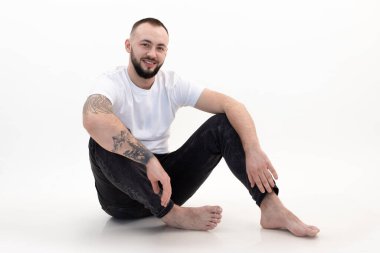 Beyaz tişörtlü, kısa siyah saçlı, sakallı genç adam, siyah kot pantolonlu, oturan, beyaz zeminde diz çökmüş..