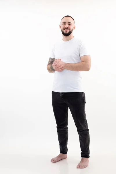 Ritratto di giovane uomo barbuto felice e sorridente con capelli corti e scuri in T-shirt bianca, jeans neri, stand che unisce le mani. — Foto Stock