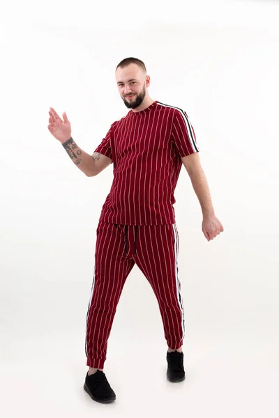 Retrato de un joven feliz hombre barbudo lindo con pelo corto y oscuro usando ropa deportiva de rayas rojas, levantando la mano, posando. —  Fotos de Stock
