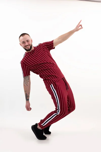 Joven hombre barbudo feliz con pelo corto y oscuro, tatuaje en ropa deportiva de rayas rojas, zapatillas negras, posando de pie en los dedos de los pies. —  Fotos de Stock