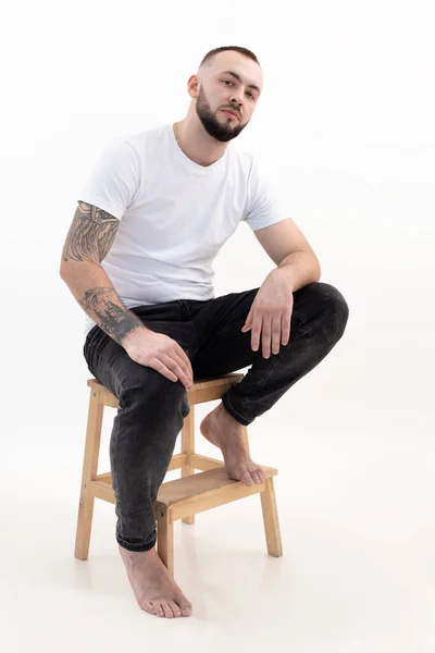Giovane bell'uomo barbuto con i capelli corti scuri in t-shirt bianca, jeans neri, sedersi sulla scala mettendo piede sulla scala. — Foto Stock