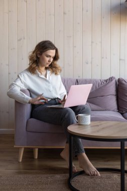 Çıplak ayaklı bir kadının portresi laptopta çalışırken yumuşak menekşe koltukta, masanın yanındaki gündelik kıyafetlerle bir fincan kahveyle oturuyor. Estetik dekorlu zarif tasarımlı oturma odası, bedava kopya alanı