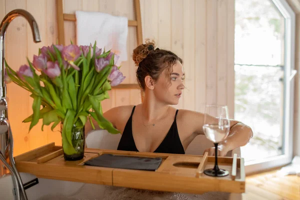 Portrait of naked woman in black underwear relaxing and spending weekend at home, soft focus. Taking hot foam bath with glass of wine on food tray and bouquet of pink tulips in cozy wooden bathroom — 스톡 사진