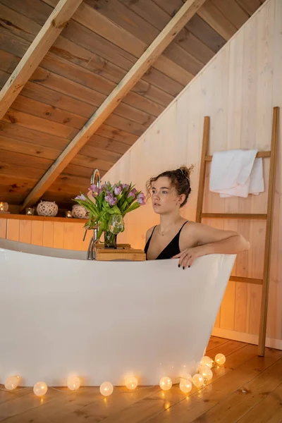 Portrait of beautiful woman in black underwear relaxing and spending weekend at home. Taking bath with glass of wine on food tray and bouquet of tulips in cozy wooden bathroom decorated with garland — 스톡 사진