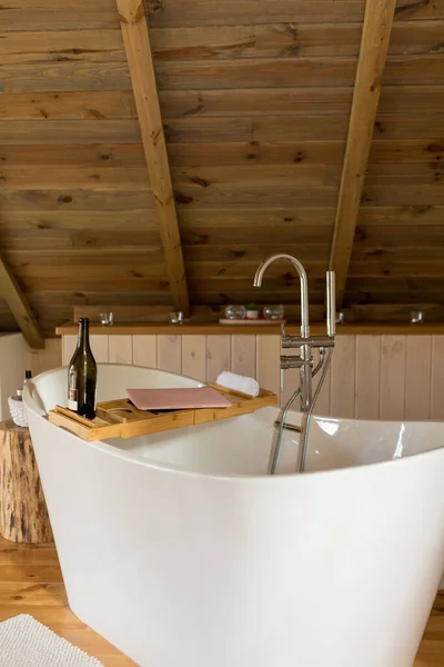 Modern wooden light bathroom interior with bathtub, water tap, bottle of wine and pink laptop on tray. Stylish bathroom accessories create comfort and coziness. Home interior and fashion decor — ストック写真