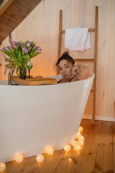 Portrait of attractive naked woman relaxing and spending weekend at home. Taking bath with glass of wine on food tray and bouquet of tulips in cozy wooden bathroom decorated with garland — 스톡 사진
