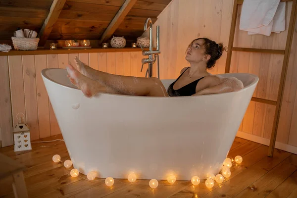 Portrait of woman in black underwear relaxing and spending weekend at home. Laying in foam bath with outstretched legs in cozy wooden bathroom decorated with garland. Home rest and bodycare concept — 스톡 사진