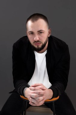 Siyah ceketli, siyah saçlı, beyaz tişörtlü, tahta taburede oturan, birbirine dolanan parmaklı, ciddi sakallı genç bir adam..