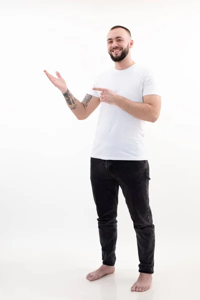Portrait de jeune homme barbu heureux aux cheveux courts et foncés en T-shirt blanc, jean noir, index pointu debout. — Photo
