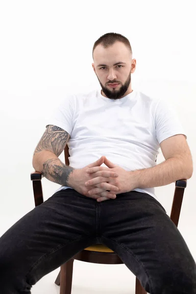 Young tranquil bearded man with short dark hair in white T-shirt, black jeans, sit on wooden chair, interlacing fingers.