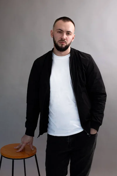 Young serious bearded man with short dark hair wearing black jacket, jeans, white T-shirt, standing at wooden stool. — Stock Photo, Image