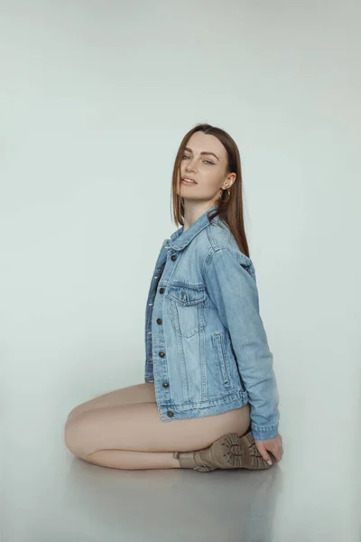 Stylish sexy woman with brown hair and perfect makeup sitting and wearing seasonal blue denim jacket and leather boots, white background. Fashion style catalog of casual clothes for young ladies —  Fotos de Stock