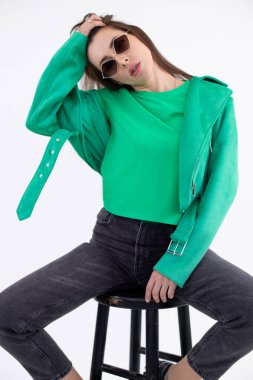 Uzun saçlı çekici bir kadının portresi yeşil tişört, ceket, gri kot pantolon, güneş gözlüğü giyer ve tabureye otururdu..