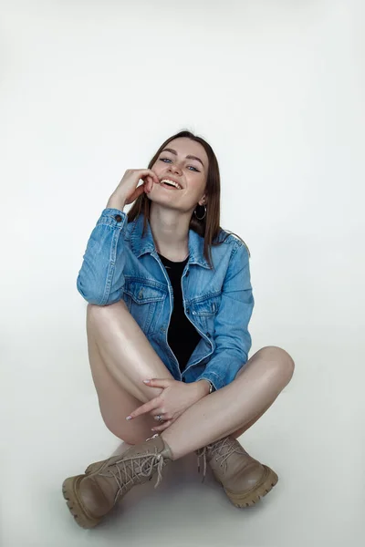 Donna calda sorridente con i capelli castani e trucco perfetto posa e indossa giacca di jeans blu stagionale e stivali di pelle, sfondo bianco. Catalogo stile moda di abiti casual per le giovani donne — Foto Stock