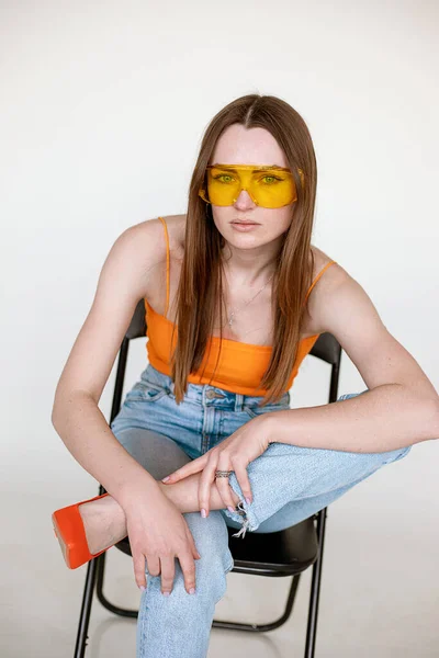 Beautiful woman in stylish sunglasses sitting on black chair and wearing seasonal blue denim and bright orange top closeup, white background. Fashion style catalog of casual clothes for young ladies — ストック写真