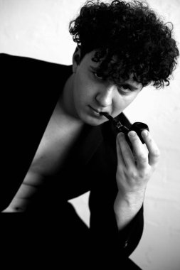 Siyah ceket giyen, kot giyen, oturan, dizine yaslanan, elinde tütün piposu tutan siyah beyaz bir adam portresi..