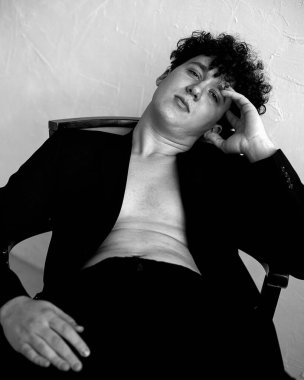 Siyah ceket giyen, kot giyen, oturan, sandalyeye yaslanan, el ele tutuşan genç adamın siyah beyaz portresi..