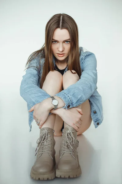Portrait of young sad woman with long dark hair, make-up wearing blue denim jacket, beige boots, sitting, hugging knees. —  Fotos de Stock