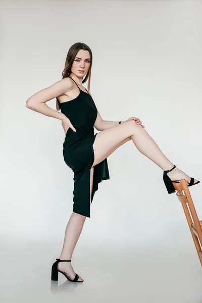 Side view of young flawless woman wearing black dress with long slit, sandals, standing, raising leg on back of chair. — Zdjęcie stockowe