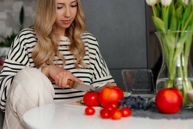 Genç, hoş bir kadın taze sebzeler kesiyor ve sandalyeye oturup sağlıklı bir yemek hazırlıyor. Modern tasarım mutfağında yemek pişiren ev elbiseli kadının portresi. Organik gıda ve düzgün beslenme