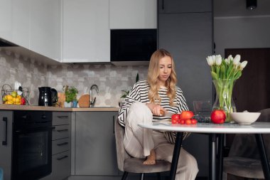 Genç, hoş bir kadın taze sebzeler kesiyor ve sandalyeye oturup sağlıklı bir yemek hazırlıyor. Modern mobilyalarla mutfakta yemek pişiren ev elbiseli kadın portresi. Organik gıda ve düzgün beslenme