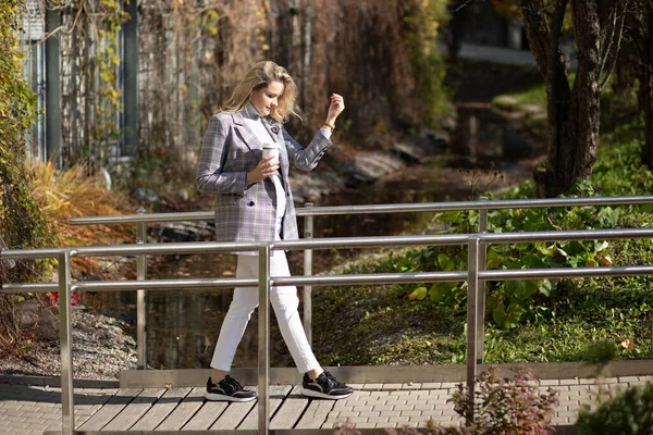 Элегантная женщина с удовольствием идет по мосту через ручей с кофе в осеннем городском парке сбоку. Бизнес-леди, гуляющая и отдыхающая на улице, городской фон. Золотая осень, выходные — стоковое фото