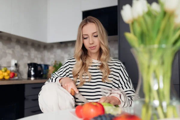 Genç, hoş bir kadın taze malzemeler kesiyor ve sandalyeye oturup sağlıklı bir yemek hazırlıyor. Modern tasarım mutfağında yemek yapan, arka planda bulanık elbiseli bir kadın. Organik gıda ve düzgün beslenme
