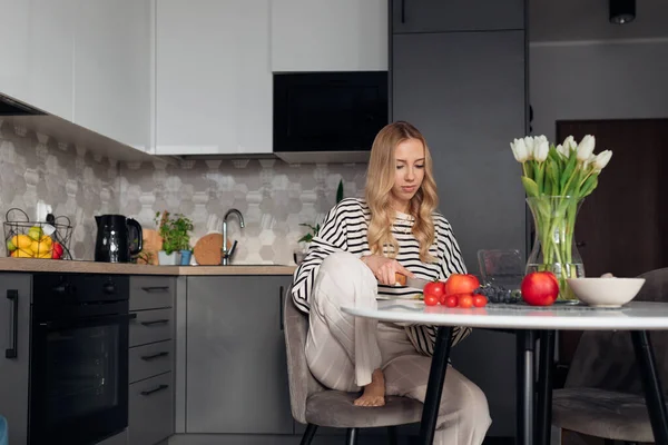Genç, hoş bir kadın taze sebzeler kesiyor ve sandalyeye oturup sağlıklı bir yemek hazırlıyor. Modern mobilyalarla mutfakta yemek pişiren ev elbiseli kadın portresi. Organik gıda ve düzgün beslenme