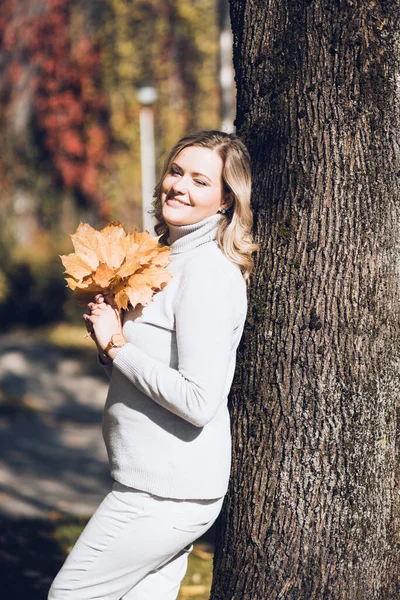 Happy woman stand leaning against tree trunk with fallen leaves in autumn city park on sunny day. Adult lady hold yellow foliage in hands and smile, nature background. Cozy golden autumn and weather — Foto Stock