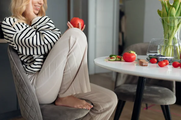 Young happy woman holding fresh red apple and smiling talking about new diet by smart phone. Portrait of lady in home clothes sitting on chair in modern kitchen. Organic food and proper nutrition — Stock Fotó
