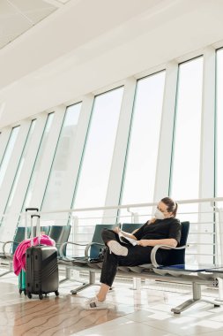 Bavulları havaalanı terminalinde oturmuş, uçuş için bekleyen, yorgun, tıbbi maskeli, sarışın bir kadının dikey hali.