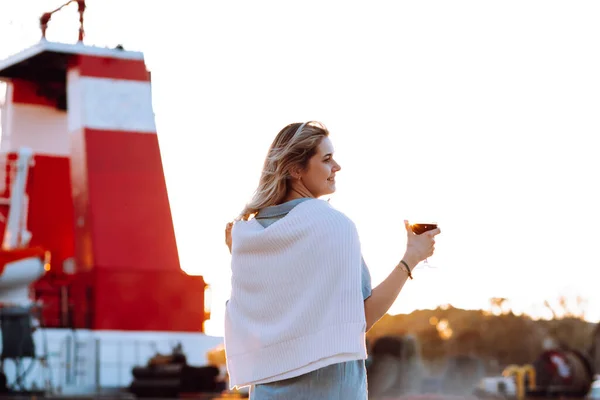 Portrait of happy woman holding in hand wine glass and drinking red wine at sunset on blurred pier background back view. Walking along sea embankment with boats and yachts in resort city