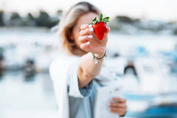 Portrait of young woman holding in hand ripe tasty strawberry on blurred background closeup, walking along embankment with boats and yachts in resort city. Sea voyage, boat mooring, relaxing leisure