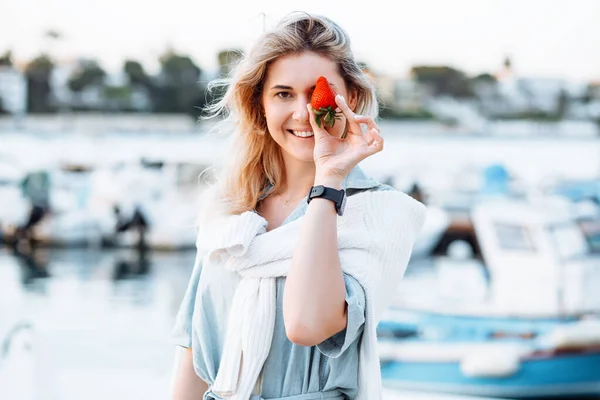 Portrait of young smiling woman holding in hand ripe tasty strawberry on blurred background, walking along embankment with boats and yachts in resort city. Sea voyage, boat mooring, relaxing leisure