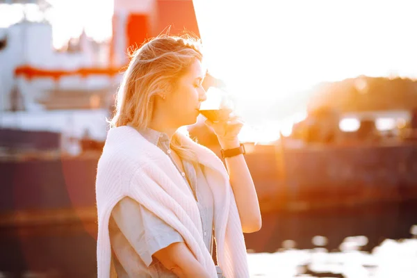 Portrait of relaxing woman holding in hand wine glass and drinking red wine at yellow sunset on blurred evening pier background. Walking along sea embankment with boats and yachts in resort city — Stock Photo, Image