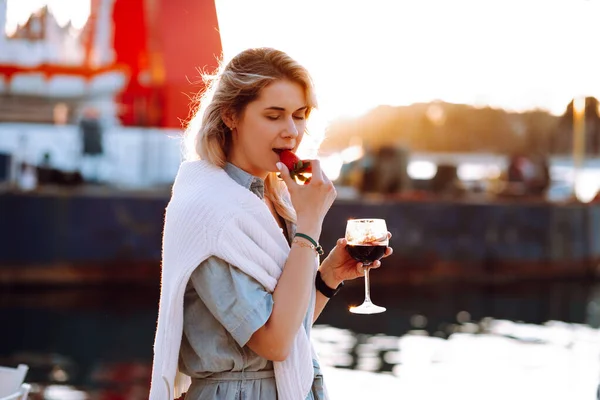 Portrait of relaxing woman holding in hand red wine glass and eating fresh strawberry at sunset on blurred evening pier background. Walking along sea embankment with boats and yachts in resort city