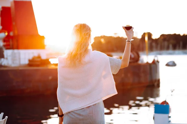 Portrait of woman holding in hand wine glass and drinking red wine at sunset on blurred evening pier background back view. Walking along sea embankment with boats and yachts in resort city