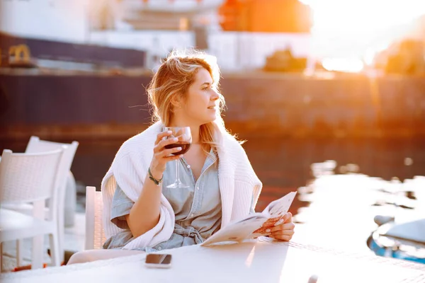 Portrait of happy woman holding in hand red wine glass and sitting at table in cafe at yellow sunset on blurred evening pier background. Walking along embankment with boats and yachts in resort city