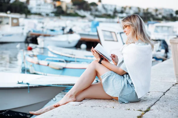 Portrait of young woman walking along embankment with boats and expensive yachts in resort city, sitting on edge of pier and reading book side view. Sea voyage, boat mooring, relaxing leisure